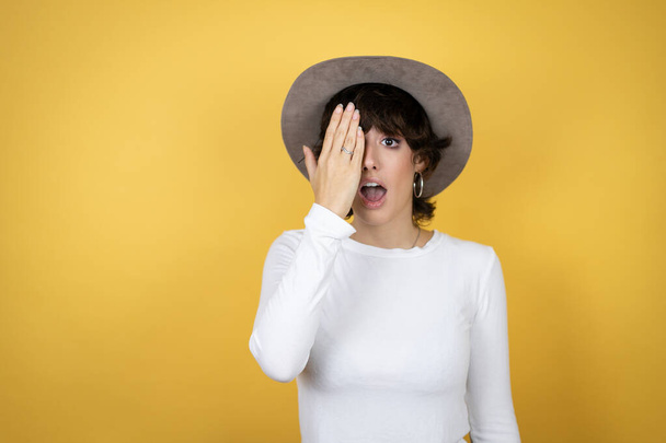 Young caucasian woman wearing hat over isolated yellow background covering one eye with hand, confident smile on face and surprise emotion. - 写真・画像