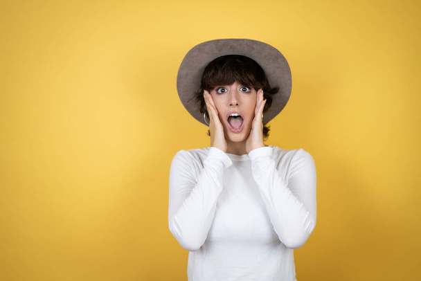 Mulher branca jovem usando chapéu sobre fundo amarelo isolado louco e assustado com as mãos na cabeça, com medo e surpreso de choque - Foto, Imagem