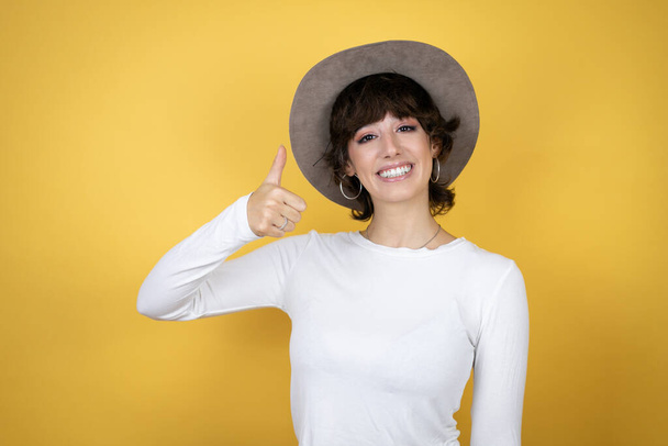 Junge kaukasische Frau mit Hut vor isoliertem gelben Hintergrund lächelt und macht das Okay-Signal mit dem Daumen - Foto, Bild