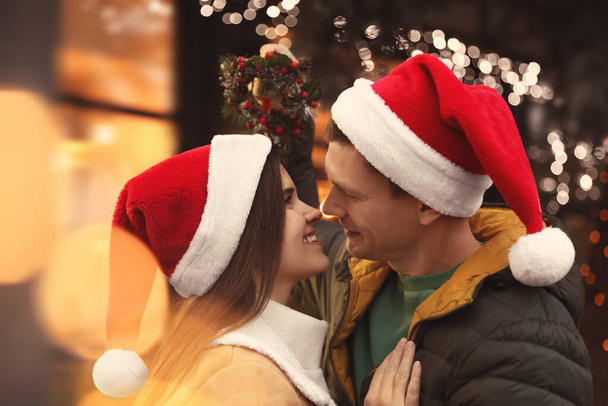 Happy couple in Santa hats standing under mistletoe wreath outdoors, bokeh effect - Фото, зображення