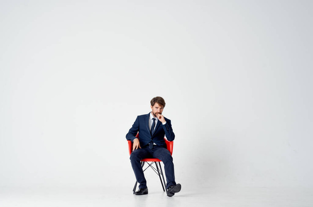 Ένας κομψός κύριος με κοστούμι κάθεται σε μια κόκκινη καρέκλα στη μέση του δωματίου. - Φωτογραφία, εικόνα