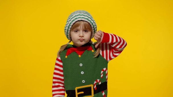 Displesed lapsi tyttö jouluna tonttu Santa auttaja puku pitää peukalo alas ja osoittaa vastenmielisyyttä ele - Valokuva, kuva