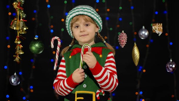 Kid girl in Christmas elf Santa Claus helper costume licking candy cane lollipop caramel sweets - Photo, Image