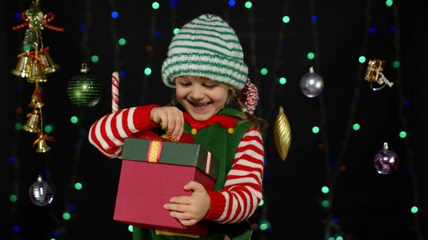 Karácsonyi manó jelmezes kislány ajándékdobozzal, aki belenéz. Kellemes ünnepeket! - Fotó, kép