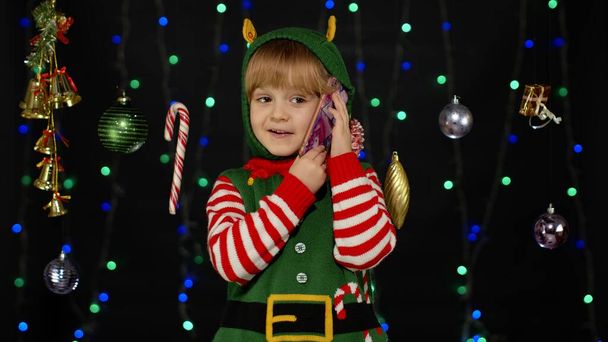 Kid girl in Christmas elf Santa Claus helper costume making congratulations call on mobile phone - Photo, Image