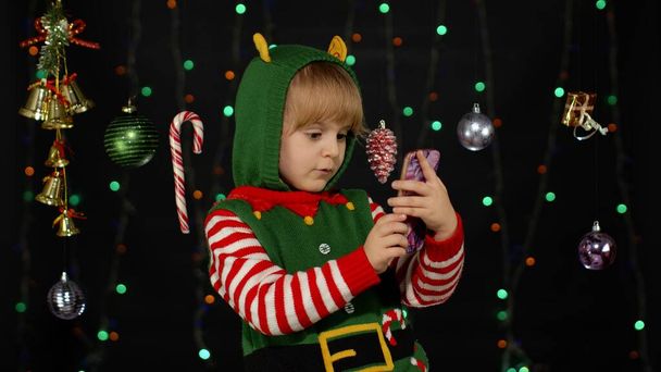 Kid girl Christmas elf Santa helper types something on mobile phone enjoys browsing social media - Photo, Image