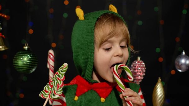 Παιδί κοριτσάκι στα Χριστούγεννα ξωτικό Άγιος Βασίλης κοστούμι βοηθός γλείφει καραμέλα γλειφιτζούρι καραμέλα γλυκά - Φωτογραφία, εικόνα