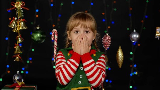 Kid girl in Christmas elf Santa Claus helper costume looks at camera makes big eyes covering mouth - Photo, Image