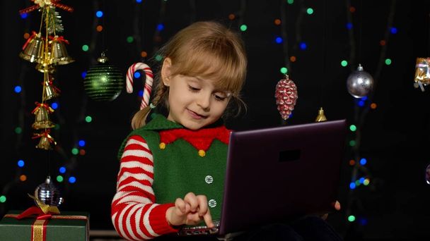 Kid girl in Christmas elf costume doing shopping online using laptop, browsing on social media - Photo, Image