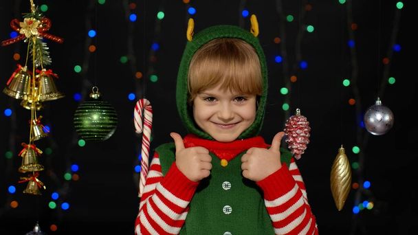 Teen kid girl in Christmas elf Santa Claus helper costume showing thumbs up on black background - Photo, Image