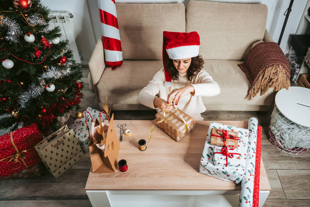 A beautiful woman preparing Christmas presents in the living room on Christmas holidays. She is using a ribbon with a gift on the table. Christmas gifts preparation concept - 写真・画像