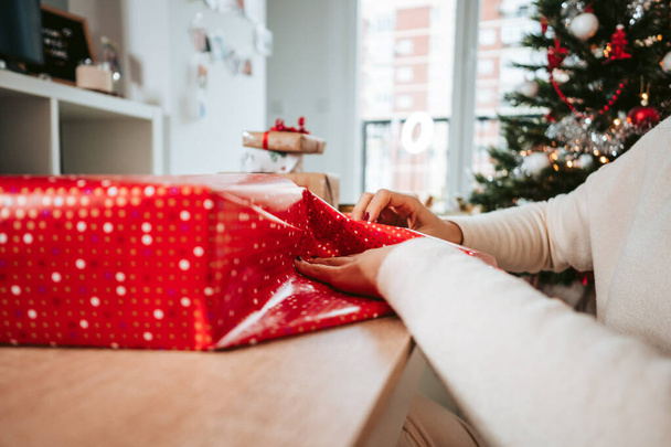 Enfoque cercano y selectivo. Una mujer que envuelve un regalo de Navidad con papel rojo en la mesa de la sala de estar. Concepto de preparación de regalos de Navidad - Foto, Imagen
