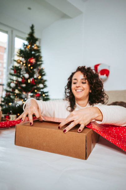 Selective focus. A brunette woman wrapping Christmas gifts in the living room whit a big Christmas tree behind her. Christmas gifts preparation concept - Фото, зображення