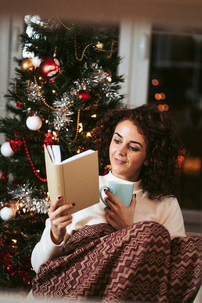 A beautiful woman reading a book in a cold day on December. She is enjoying a cup of tea while she is covering herself with a blanket. Christmas holidays concept - Foto, Imagen