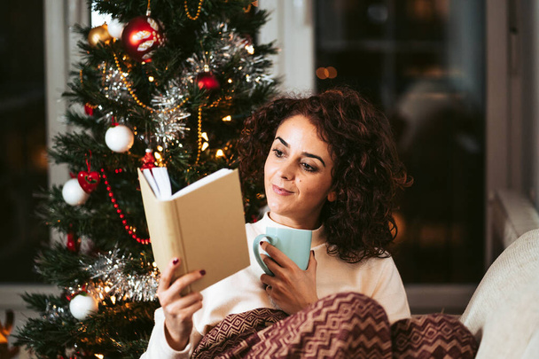A brunette woman sitting on the sofa while reading a book in a cold day on December. She is near to a Christmas tree and she is covering herself with a blanket. Christmas holidays concept. - Photo, image