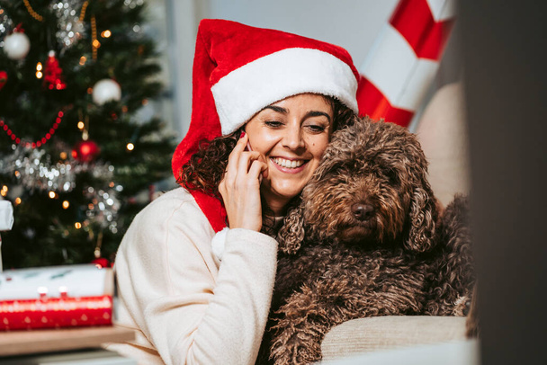 Μια όμορφη γυναίκα που διασκεδάζει με τον Ισπανό σκύλο της ενώ μιλάει στο τηλέφωνο. Φοράει καπέλο του Άγιου Βασίλη. Οικογενειακό σκυλί Χριστουγεννιάτικο δέντρο έννοια - Φωτογραφία, εικόνα