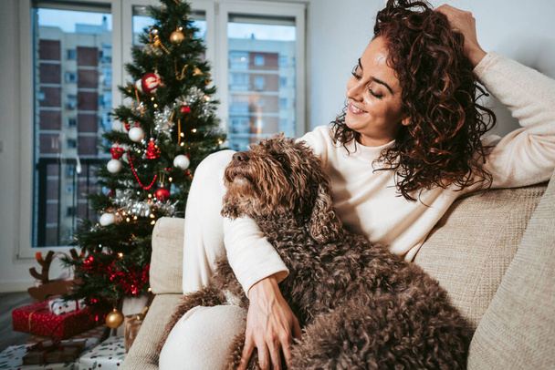 brunetka sedící na gauči se svým španělským vodním psem a dívající se na něj s láskou na Štědrý den. Jsou uvolněné o vánočních svátcích. Vánoční rodinný pes koncept - Fotografie, Obrázek