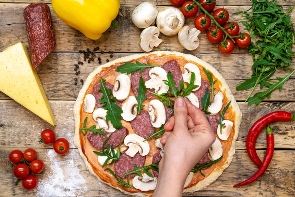 ingredientes para hacer pizza, antes de hornear, en una mesa de madera, vista superior de la mano expuesta rúcula verdes, receta paso a paso. Foto de alta calidad - Foto, imagen
