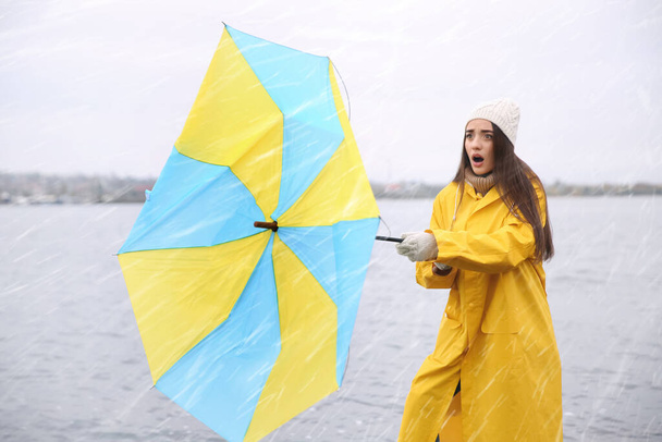 Woman in yellow raincoat with umbrella caught in gust of wind near river - Foto, imagen