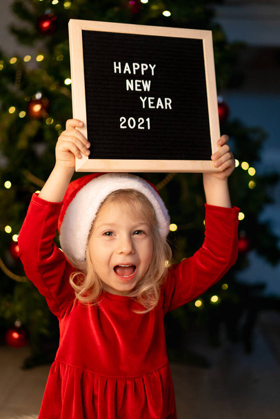 Noel Baba şapkası ve kırmızı Noel elbisesi ile küçük güzel kız Noel ağacının altında yatıyor ve gülüyor, siyah bir tahtanın yanında ve beyaz harflerle mutlu yıllar 2021 mesajı atıyor. Yüksek kalite fotoğraf - Fotoğraf, Görsel