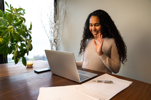 Beautiful young mixed race African American woman waves her hand to say hello and smiles on a video conference call on her laptop at a temporary remote work from home station at her dining table. - Foto, imagen