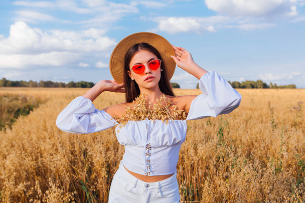 Rural Countryside Scene. Fashion portrait of a young beautiful woman with long hair dressed in white blouse, straw hat and pink sunglasses at golden oat field with ears of oats - Valokuva, kuva