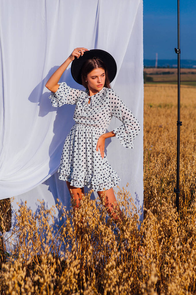 Rural Countryside Scene. Young beautiful woman with long hair dressed in white Polka-dot dress and black hat standing at golden oat field with a white curtain on the background. - Фото, изображение