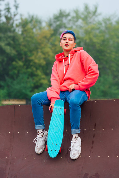 Summer lifestyle image of trendy pretty young girl with short colored hair and nose piercing dressed in pink hoodie and jeans sitting next to the skateboard court with her blue plastic skateboard. - Photo, Image