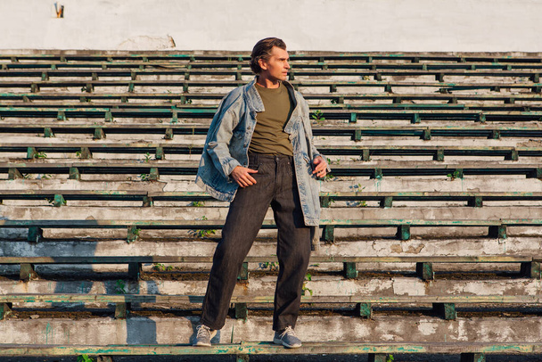 Fashion portrait of a tall handsome man dressed in jeans jacket on the crushed tribune of the old stadium during sunset - 写真・画像