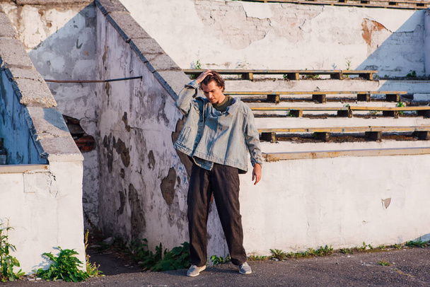 Fashion portrait of a tall handsome man dressed in jeans jacket on the crushed tribune of the old stadium during sunset - Valokuva, kuva