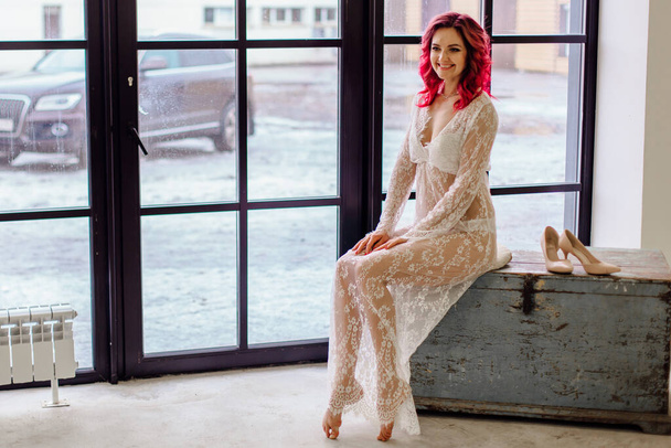Attractive woman with pink hair in white light dress sitting on the old wooden box near large window. Lady in gentle lace peignoir sitting with bare feet in a room with concrete floor - Zdjęcie, obraz