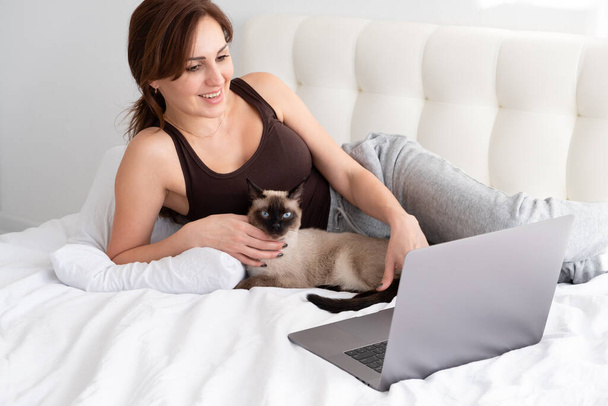 Alkalmi nő laptopot használ a sziámi macskájával a fehér hálószobában. kedvtelésből tartott állatok. - Fotó, kép