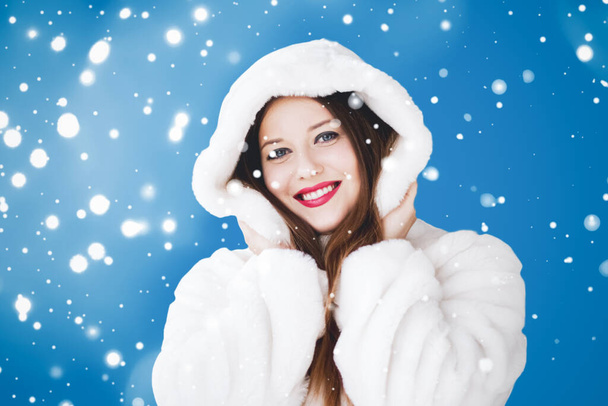 Happy Christmas en winter vakantie portret van jonge vrouw in witte kap bontjas, sneeuw op blauwe achtergrond, mode en levensstijl - Foto, afbeelding