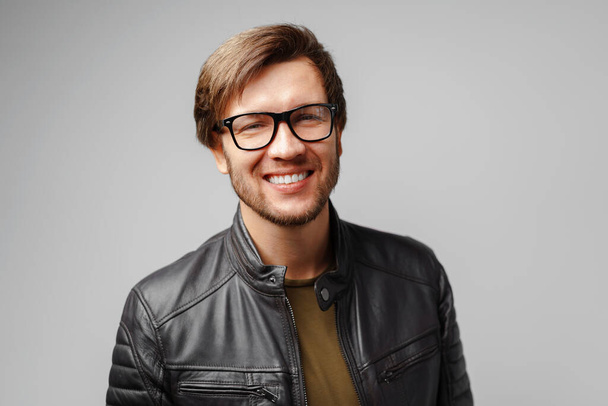 Portrait of a young man in glasses wearing black leather jacket on grey background - Foto, imagen