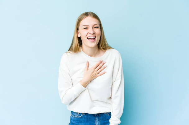 Young blonde woman isolated on blue background laughs out loudly keeping hand on chest. - Foto, immagini