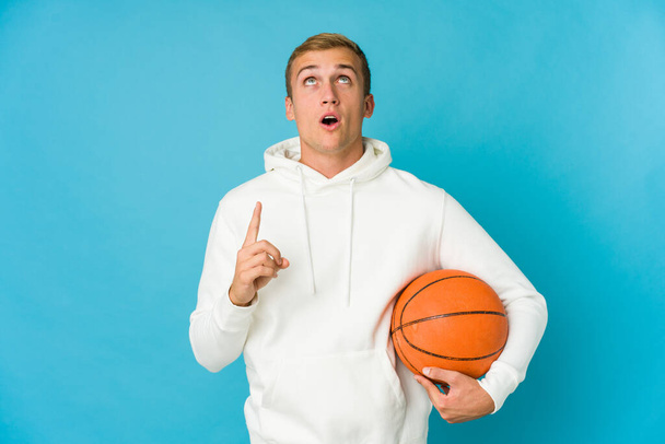 jonge Kaukasische man spelen basketbal geïsoleerd op blauwe achtergrond wijzend ondersteboven met geopende mond. - Foto, afbeelding