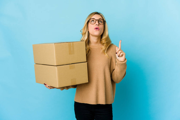 Молодая блондинка, белая женщина, держит коробки, чтобы двигаться, указывая вверх, с открытым ртом. - Фото, изображение