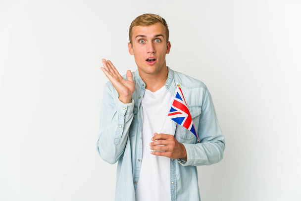 Beyaz arka planda izole edilmiş Birleşik Krallık bayrağı tutan genç beyaz adam şaşırdı ve şok oldu.. - Fotoğraf, Görsel
