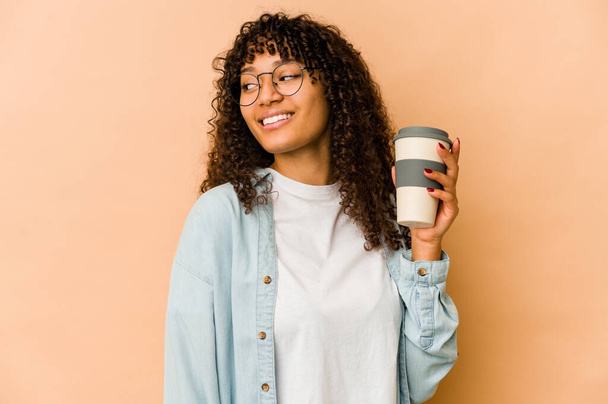 Joven afroamericana afro mujer sosteniendo un café para llevar mira a un lado sonriente, alegre y agradable. - Foto, Imagen