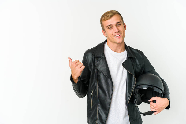 Joven hombre caucásico sosteniendo un casco de moto aislado en puntos de fondo blanco con el dedo pulgar lejos, riendo y despreocupado. - Foto, imagen