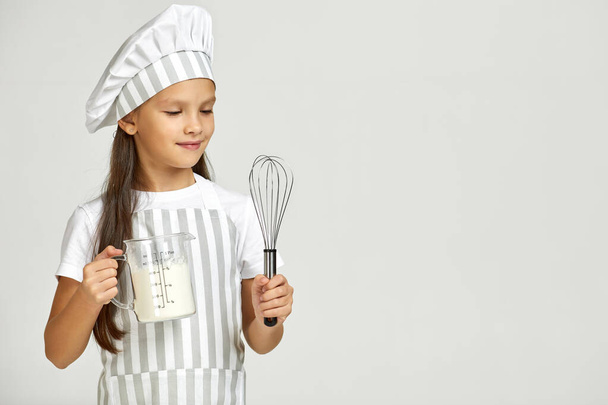 Aşçı şapkalı ve önlüklü küçük kız ölçü kabında un ve çırpı. - Fotoğraf, Görsel