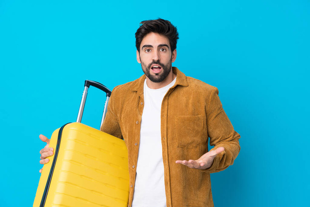 Młody przystojny mężczyzna z brodą na odizolowanym niebieskim tle na wakacjach z walizką podróżną - Zdjęcie, obraz