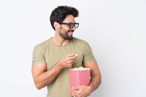 Junger gutaussehender Mann mit Bart auf isoliertem weißem Hintergrund mit 3D-Brille und einem großen Eimer Popcorn - Foto, Bild