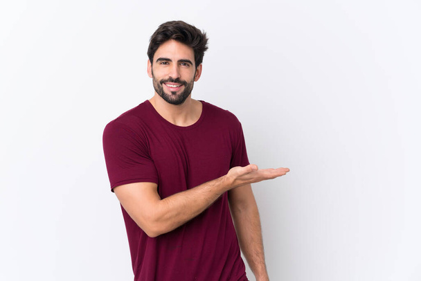 Young handsome man with beard over isolated white background presenting an idea while looking smiling towards - Photo, Image