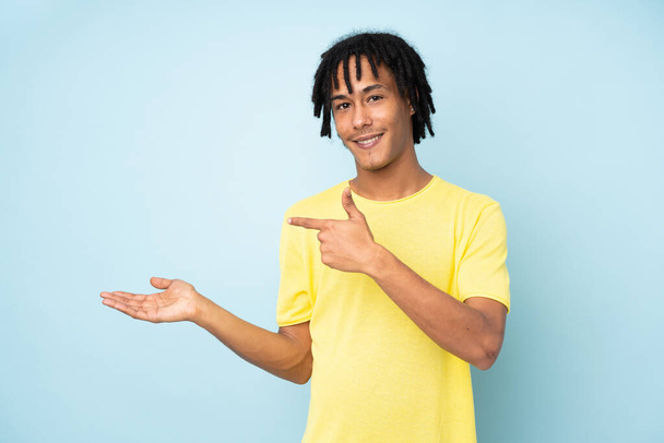 Jonge Afro-Amerikaanse man geïsoleerd op blauwe achtergrond houden copyspace imaginaire op de palm om een advertentie in te voegen - Foto, afbeelding