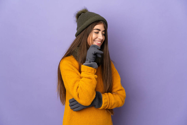 Mladý kavkazský dívka s zimní klobouk izolované na fialovém pozadí dívá do strany a usmívá - Fotografie, Obrázek