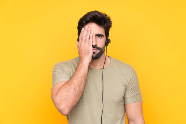 Telemarketer muž pracuje se sluchátky přes izolované žluté pozadí zakrývající oko po ruce - Fotografie, Obrázek