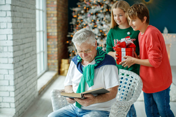 Grootvader en kleinkinderen in een kamer versierd voor Kerstmis tegen de achtergrond van een kerstboom. Kerstvakantie concept. contrastfotografie. - Foto, afbeelding