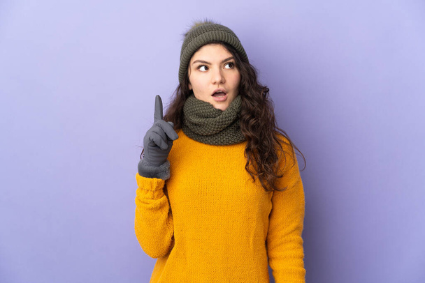 Έφηβη Ρωσίδα με χειμερινό καπέλο που απομονώθηκε σε μωβ φόντο σκεπτόμενη μια ιδέα που δείχνει το δάχτυλο προς τα πάνω - Φωτογραφία, εικόνα