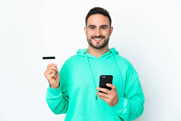 Jonge blanke knappe man geïsoleerd op witte achtergrond kopen met de mobiele telefoon met een creditcard - Foto, afbeelding